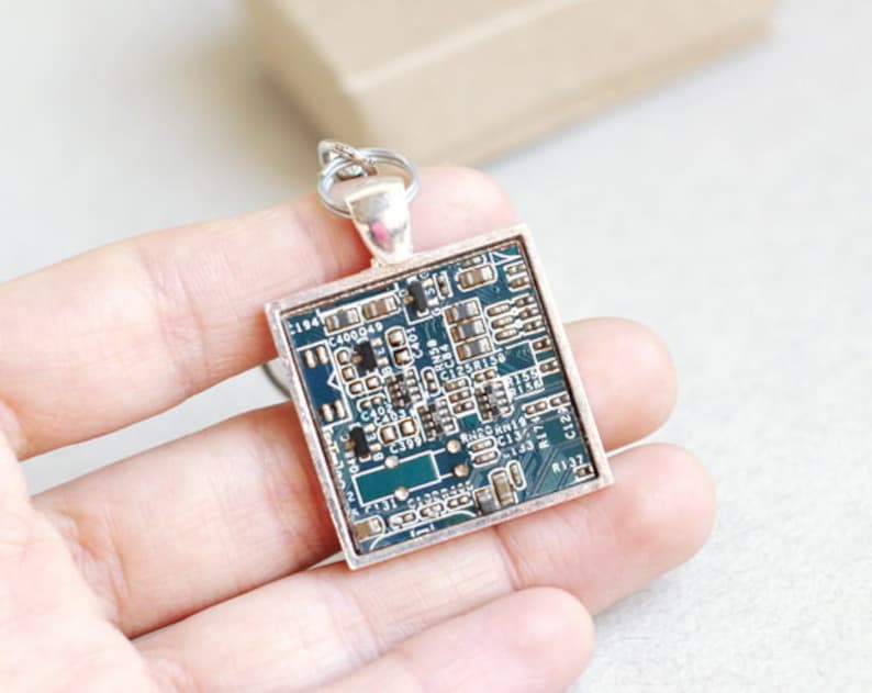 May include: Square pendant featuring a section of a circuit board encased in a silver-toned frame. The circuit board has a teal background with various electronic components. The pendant is attached to a silver-toned bail.