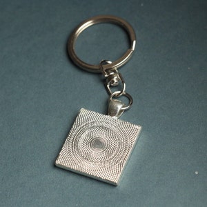 May include: Silver-toned keychain with a square pendant. The pendant features a detailed, textured design with a central circular pattern. The keychain includes a split ring and a lobster clasp for attaching keys or accessories.