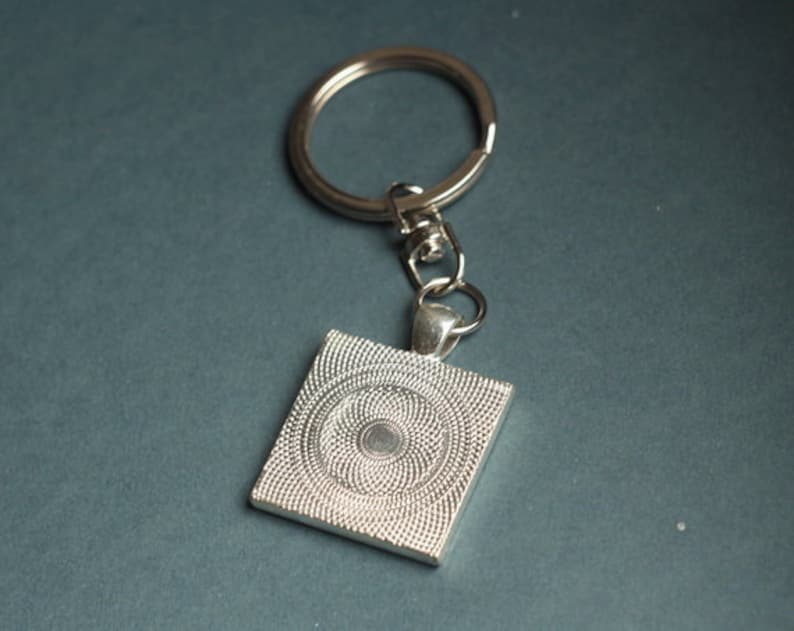 May include: Silver-toned keychain with a square pendant. The pendant features a detailed, textured design with a central circular pattern. The keychain includes a split ring and a lobster clasp for attaching keys or accessories.
