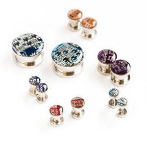 Può includere: Una collezione di plug e orecchini a perno con design a circuito elettronico. I plug hanno una base argentata e un design colorato a circuito stampato in blu, rosso, arancione e viola. Gli orecchini hanno un design simile.