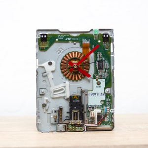 Puede incluir: Un reloj hecho a partir de un disco duro de computadora reutilizado con una aguja de segundos roja y una esfera de color cobre. El disco duro es verde y plateado con placas de circuito visibles y texto que dice "2X12A06F".