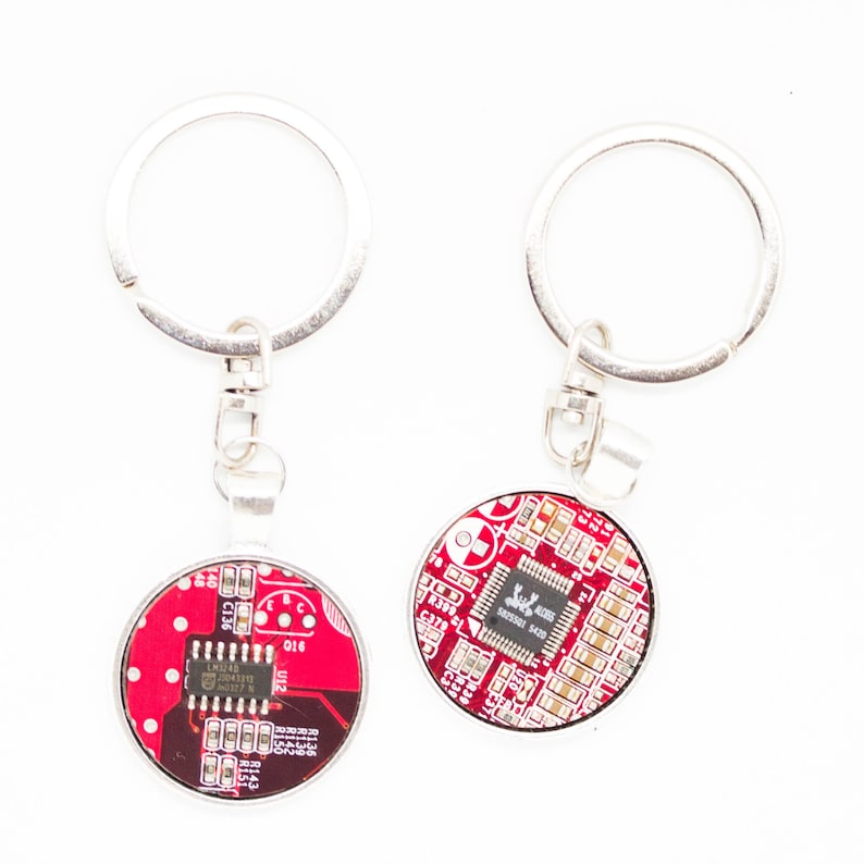 May include: Two silver keychains with round, red circuit board designs. Each keychain has a silver ring and a clasp. The circuit boards feature various electronic components and text. The keychains are displayed against a white background.