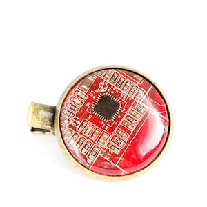 Pode incluir: Um pingente redondo vermelho e dourado com um design de placa de circuito. A placa de circuito está encapsulada em resina transparente e tem um laço de metal dourado para prender uma corrente.