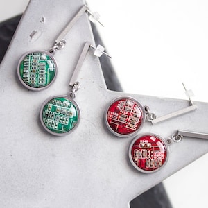May include: Three pairs of earrings with circular green and red circuit board designs. The earrings are silver and have a long, straight post.