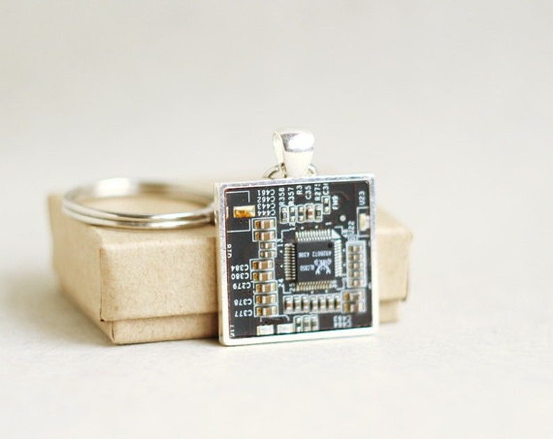 May include: A square silver-toned keychain pendant featuring a detailed circuit board design. The pendant is black with gold and white accents, attached to a silver keyring. The pendant is resting on a small, brown cardboard box.