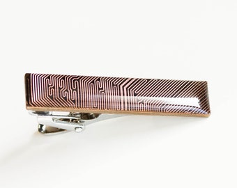 May include: A copper-coloured tie clip featuring a circuit board design. The rectangular clip has a shiny, metallic finish and a detailed pattern of lines and shapes. The clip is set against a white background.