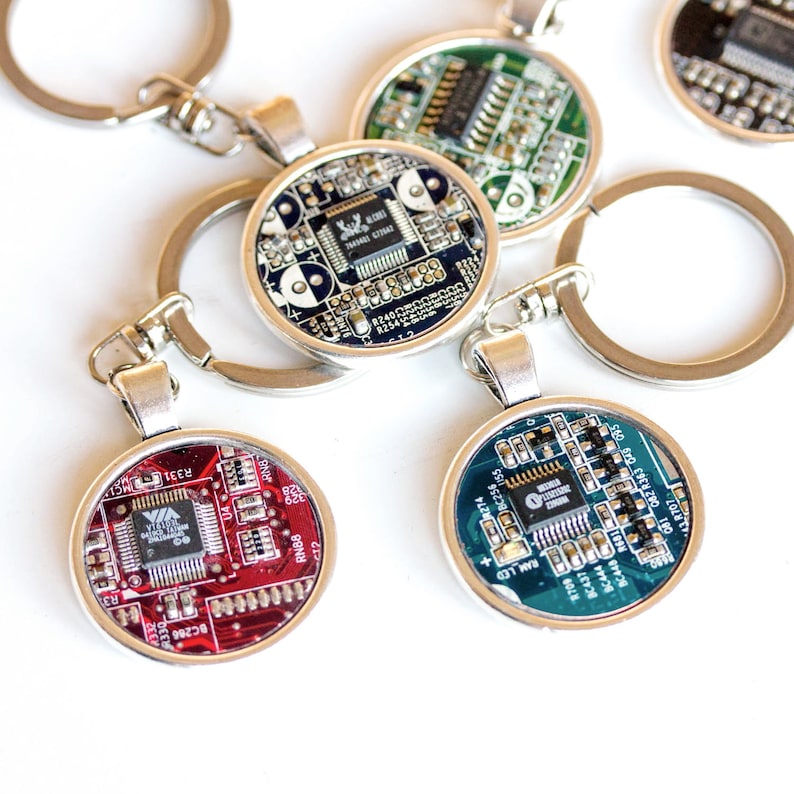May include: A collection of round keychains featuring circuit board designs in various colors, including red, green, and blue. Each keychain has a silver-colored metal frame and a key ring. The circuit board components are visible within the clear resin.