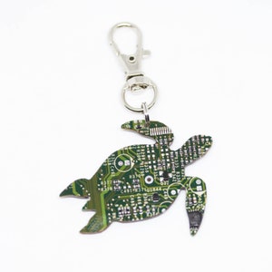 Peut inclure: Un porte-clés en forme de tortue fabriqué à partir d'une carte de circuit imprimé verte. Le porte-clés est doté d'un fermoir argenté. Le design de la carte de circuit imprimé présente de petits composants et du texte.