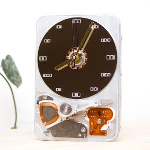 Puede incluir: Un reloj único hecho de un disco duro de computadora reutilizado. La esfera del reloj es un círculo marrón oscuro con números binarios blancos que marcan las horas. Las manecillas doradas indican la hora. La carcasa metálica plateada del reloj revela componentes internos.