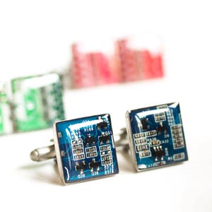 May include: A pair of cufflinks featuring a blue circuit board design. The cufflinks are square shaped and have silver metal accents.