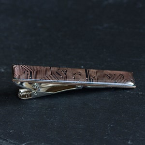 Könnte beinhalten: Ein silberner Krawattenclip mit einem schwarzen und kupferfarbenen Leiterplatten-Design.