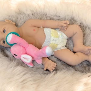 May include: A baby doll with blue eyes and a pacifier is lying on a fluffy, gray and white faux fur blanket. The doll is wearing a diaper and holding a pink stuffed bunny. The doll's skin is a light peach color.