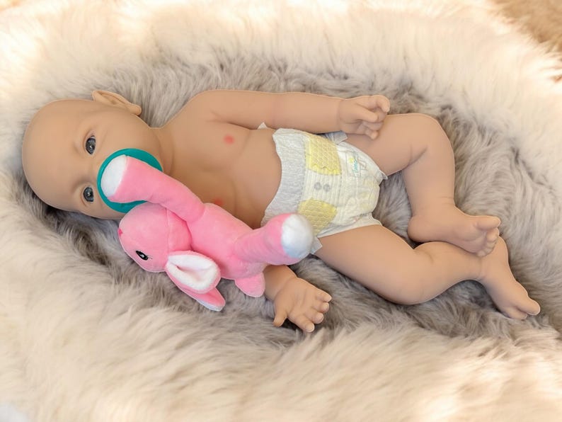 May include: A baby doll with blue eyes and a pacifier is lying on a fluffy, gray and white faux fur blanket. The doll is wearing a diaper and holding a pink stuffed bunny. The doll's skin is a light peach color.