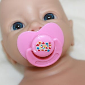 May include: A close-up of a baby doll with blue eyes and a pink pacifier. The pacifier has a round center with the words "Happy Baby" and colorful star and heart designs.
