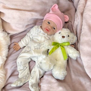 May include: A baby doll wearing a white onesie with a gray pattern and a pink hat with ears, lying on a pink blanket. A white teddy bear with a green ribbon is next to the doll.