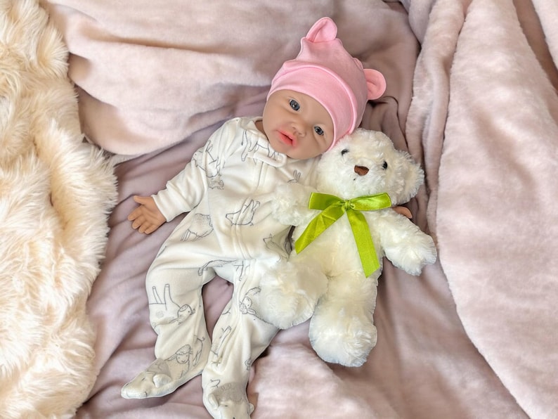 May include: A baby doll wearing a white onesie with a gray pattern and a pink hat with ears, lying on a pink blanket. A white teddy bear with a green ribbon is next to the doll.