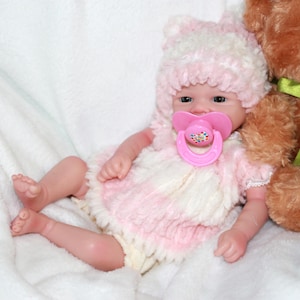 A baby doll with a pink hat and a pacifier, resting on a soft surface, showcasing its cute features.