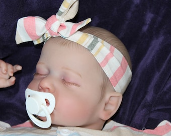 Puede incluir: Una muñeca bebé durmiendo con una diadema a rayas rosa y blanca y un chupete blanco. La muñeca tiene rasgos realistas y lleva un atuendo blanco y rosa.