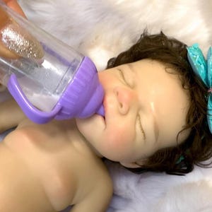 Op de afbeelding: Een babypop met gesloten ogen wordt gevoed uit een doorzichtige plastic fles met een paarse handgreep en speen. De pop heeft donker haar en een turquoise libelle haaraccessoire. De pop ligt op een witte, zachte ondergrond.