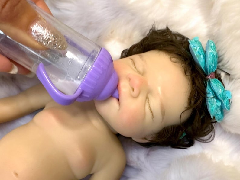 Op de afbeelding: Een babypop met gesloten ogen wordt gevoed uit een doorzichtige plastic fles met een paarse handgreep en speen. De pop heeft donker haar en een turquoise libelle haaraccessoire. De pop ligt op een witte, zachte ondergrond.
