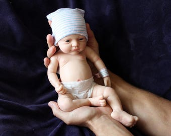 Op de afbeelding: Een realistische babypop met een lichte huidskleur, met een gestreepte blauw-witte hoed en een luier. De pop wordt in iemands handen gehouden tegen een donkerblauwe achtergrond. De pop heeft een ziekenhuisarmband.