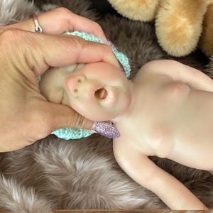 Op de afbeelding: Een realistische babypop met een lichte huidskleur, met een kleine, lichtblauwe gehaakte muts. De pop ligt op een zacht, harig oppervlak. De mond van de pop staat open. Een hand met een ring en glitterende nagels houdt het hoofd van de pop vast.