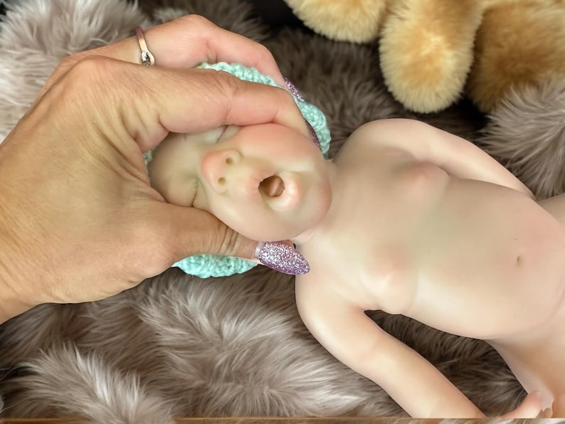 Op de afbeelding: Een realistische babypop met een lichte huidskleur, met een kleine, lichtblauwe gehaakte muts. De pop ligt op een zacht, harig oppervlak. De mond van de pop staat open. Een hand met een ring en glitterende nagels houdt het hoofd van de pop vast.