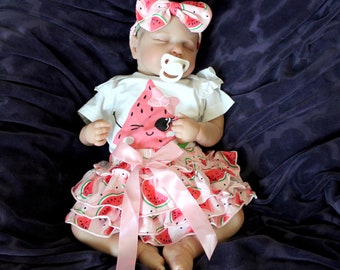 May include: A baby doll wearing a white onesie with a watermelon graphic and a pink ruffled skirt with a watermelon print. The doll is wearing a pink headband with a watermelon print and has a pacifier in its mouth.
