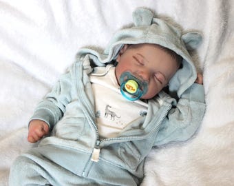 Puede incluir: Un muñeco bebé durmiente con un mono con capucha azul claro con orejas de oso y una chaqueta con cremallera a juego. El muñeco tiene un chupete y está acostado sobre una manta blanca. El mono tiene una camisa blanca debajo con un gráfico de animal gris.