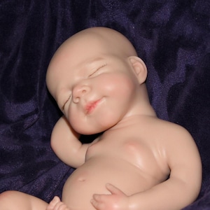 Op de afbeelding: Een levensechte babypop met gesloten ogen en een serene uitdrukking. De pop heeft een gladde, bleke huidskleur en ligt op een donkerpaarse stof. De lippen van de pop zijn lichtjes geopend en haar armen zijn zachtjes gebogen.