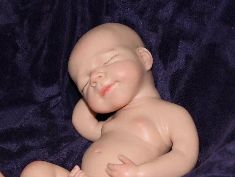 Op de afbeelding: Een levensechte babypop met gesloten ogen en een serene uitdrukking. De pop heeft een gladde, bleke huidskleur en ligt op een donkerpaarse stof. De lippen van de pop zijn lichtjes geopend en haar armen zijn zachtjes gebogen.