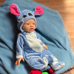 May include: A baby doll dressed in a blue jumpsuit with a hood featuring large, pink-lined ears. The doll is lying on a blue blanket. The jumpsuit has a light blue belly and feet, and a zipper closure.