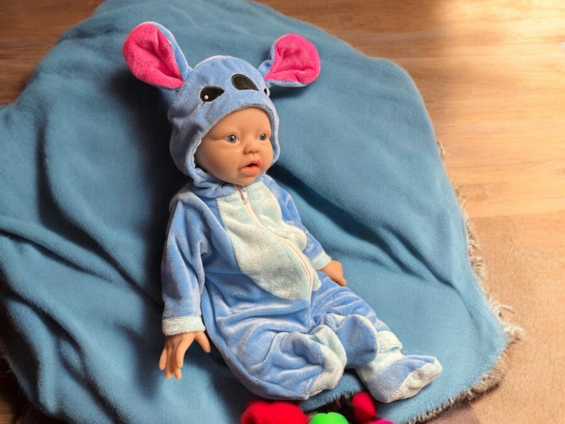May include: A baby doll dressed in a blue jumpsuit with a hood featuring large, pink-lined ears. The doll is lying on a blue blanket. The jumpsuit has a light blue belly and feet, and a zipper closure.
