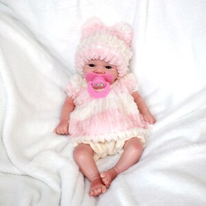 A baby doll with a pink hat and a pacifier, resting on a soft surface, showcasing its cute features.