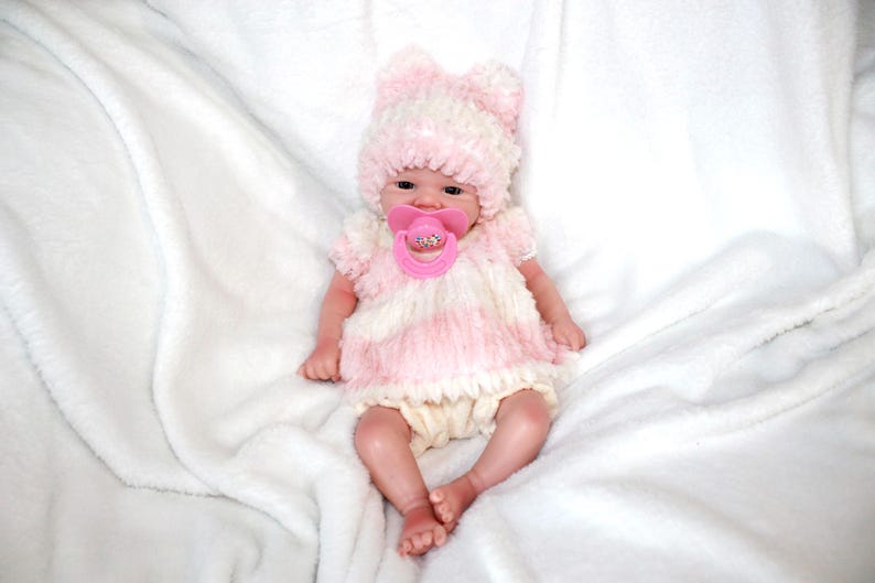 A baby doll with a pink hat and a pacifier, resting on a soft surface, showcasing its cute features.