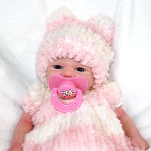 A baby doll with a pink hat and a pacifier, resting on a soft surface, showcasing its cute features.