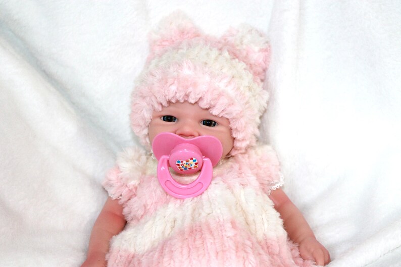 A baby doll with a pink hat and a pacifier, resting on a soft surface, showcasing its cute features.