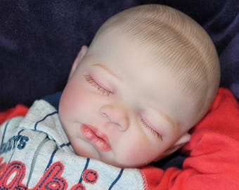 Puede incluir: Una muñeca bebé durmiendo con piel clara, ojos cerrados y una expresión serena. La muñeca tiene cabello castaño claro y lleva una camisa a rayas grises y rojas. El fondo es de color azul oscuro.