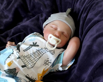 Puede incluir: Un muñeco de bebé durmiendo que lleva un gorro gris y un mono blanco con estampado de dinosaurio. El muñeco tiene un chupete en la boca.