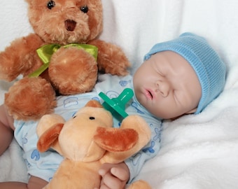 May include: A sleeping baby doll wearing a blue hat and a blue and white onesie. The doll is holding a green pacifier and a brown plush puppy. A brown teddy bear with a green ribbon is in the background.