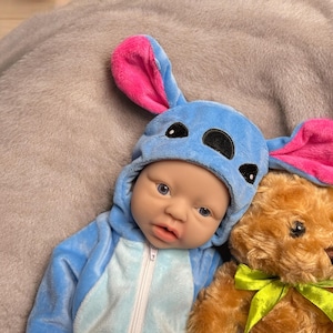 May include: A baby doll wearing a blue and white jumpsuit and a matching blue hat with pink ears. A brown teddy bear with a green ribbon is next to the doll on a gray surface.