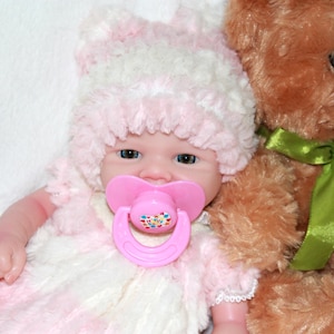 A baby doll with a pink hat and a pacifier, resting on a soft surface, showcasing its cute features.