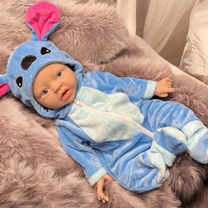 May include: A baby doll dressed in a blue and light blue plush jumpsuit with a matching hood featuring large pink and blue ears. The doll is lying on a fluffy, gray faux fur blanket.