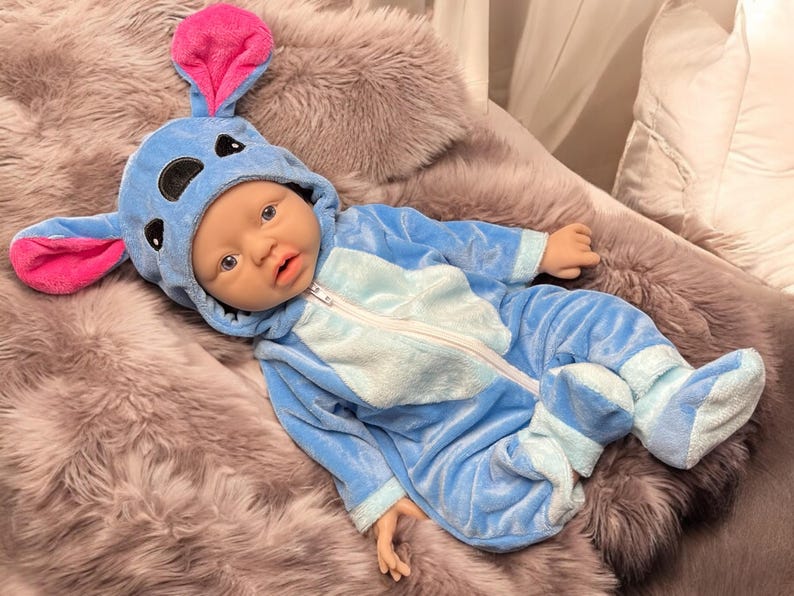 May include: A baby doll dressed in a blue and light blue plush jumpsuit with a matching hood featuring large pink and blue ears. The doll is lying on a fluffy, gray faux fur blanket.