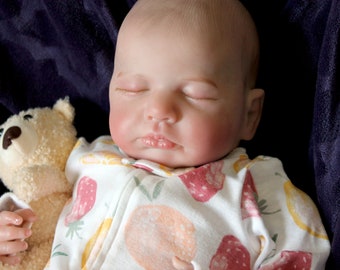 Puede incluir: Un muñeco de bebé durmiendo que lleva un mono blanco y rosa con estampado de fresas. El muñeco lleva un oso de peluche marrón.
