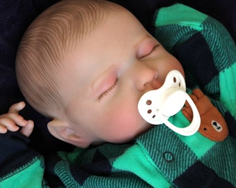 Puede incluir: Un bebé muñeca durmiendo con un mono de franela verde y negro a cuadros y un chupete blanco en la boca.