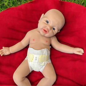 May include: A realistic baby doll with a bald head, blue eyes, and a pacifier-shaped mouth, wearing a diaper with yellow accents. The doll is lying on a red blanket, with its arms and legs spread out.