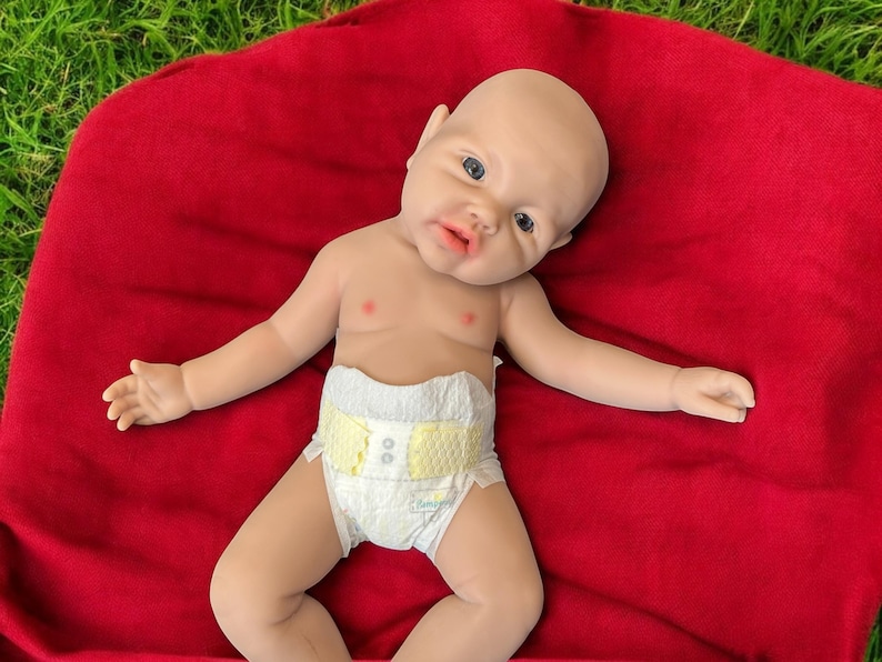 May include: A realistic baby doll with a bald head, blue eyes, and a pacifier-shaped mouth, wearing a diaper with yellow accents. The doll is lying on a red blanket, with its arms and legs spread out.