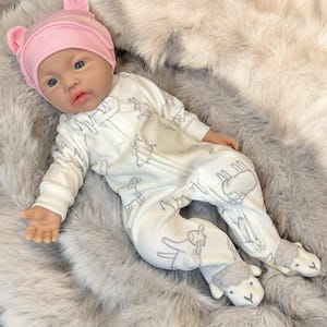 May include: A lifelike baby doll wearing a pink bear-eared hat and a white onesie with gray sheep and llama patterns. The doll is lying on a soft, gray faux fur blanket. The onesie has a zipper closure.