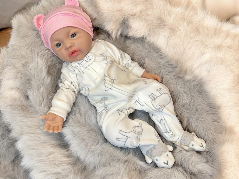 May include: A lifelike baby doll wearing a pink bear-eared hat and a white onesie with gray sheep and llama patterns. The doll is lying on a soft, gray faux fur blanket. The onesie has a zipper closure.